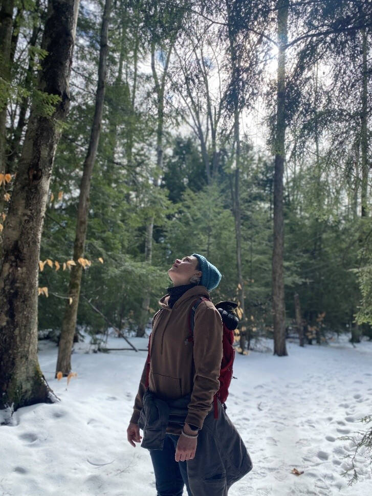 Grace Forthaus gazes up into a hemlock forest canopy from snow-covered ground.